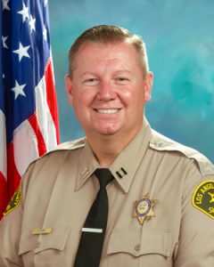Captain Vienna is wearing a tan long sleeve shirt with a black tie, badge on left side of shirt, sitting in front of a blue background, the american flag is behind him, over his right shoulder.